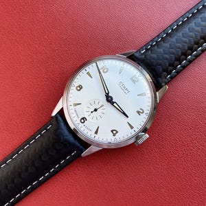 Peut inclure: Une montre-bracelet vintage avec un boîtier circulaire argenté et un cadran blanc comportant des chiffres arabes noirs à 2, 6, 8, 10 et 12. La montre comprend un sous-cadran, des aiguilles noires et un bracelet en cuir noir texturé.