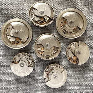 May include: Seven silver watch movements are displayed, revealing intricate internal gears and mechanisms. The circular components are arranged on a textured grey surface, highlighting the detailed craftsmanship of vintage timepieces.