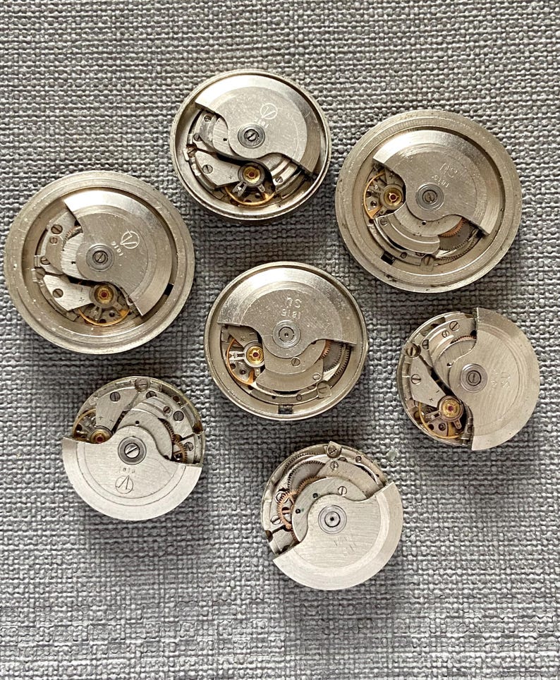 May include: Seven silver watch movements are displayed, revealing intricate internal gears and mechanisms. The circular components are arranged on a textured grey surface, highlighting the detailed craftsmanship of vintage timepieces.