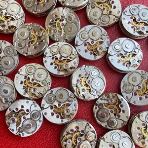 Peut inclure: Une collection de 18 mouvements de montres vintage avec des engrenages complexes et des rubis. Les mouvements sont argent&eacute;s et ont une finition dor&eacute;e. Les mouvements de montres sont dispos&eacute;s de mani&egrave;re al&eacute;atoire sur une surface rouge.