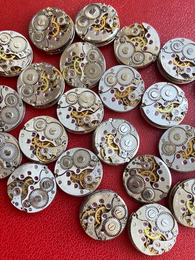 Peut inclure: Une collection de 18 mouvements de montres vintage avec des engrenages complexes et des rubis. Les mouvements sont argent&eacute;s et ont une finition dor&eacute;e. Les mouvements de montres sont dispos&eacute;s de mani&egrave;re al&eacute;atoire sur une surface rouge.