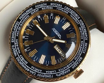 May include: A gold-toned wristwatch with a blue face and a black leather band. The watch face features a world time dial with city names in Cyrillic and Latin script. The watch face also includes a date window and a 24-hour hand.