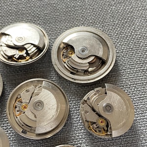 May include: Close-up of several disassembled watch movements. The silver mechanisms reveal intricate gears, springs, and other components. The watch parts are arranged on a grey surface, showcasing the detailed craftsmanship of the timepieces.