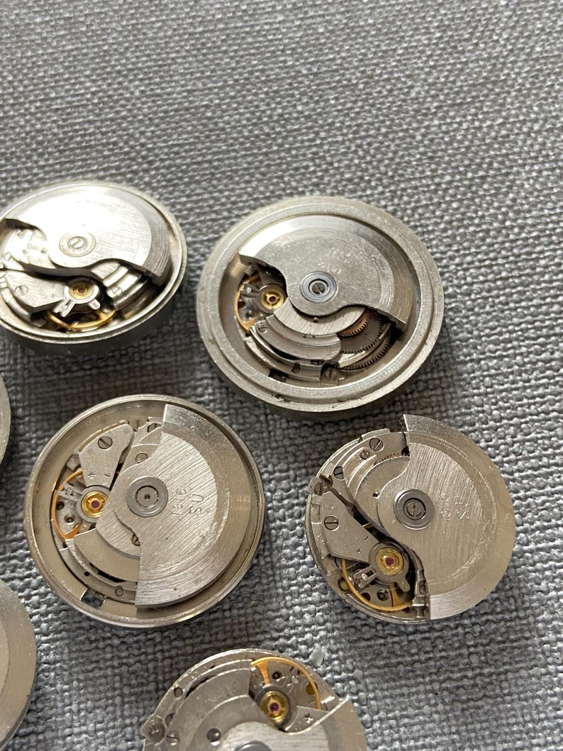 May include: Close-up of several disassembled watch movements. The silver mechanisms reveal intricate gears, springs, and other components. The watch parts are arranged on a grey surface, showcasing the detailed craftsmanship of the timepieces.