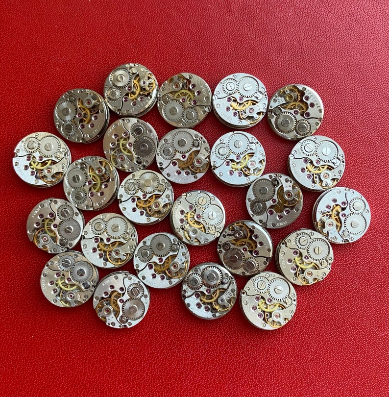 Peut inclure: Une collection de 25 mouvements de montres argent&eacute;s avec des engrenages et des ressorts complexes. Les mouvements de montres sont dispos&eacute;s en cercle sur une surface rouge.