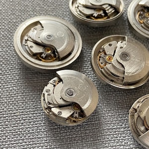 May include: Several disassembled watch movements, revealing the complex internal mechanisms. The silver-coloured components display gears, springs, and other delicate parts, demonstrating the precision engineering of timepieces. The watch parts are arranged on a textured surface.