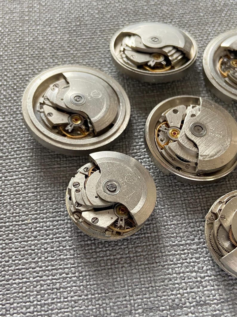 May include: Several disassembled watch movements, revealing the complex internal mechanisms. The silver-coloured components display gears, springs, and other delicate parts, demonstrating the precision engineering of timepieces. The watch parts are arranged on a textured surface.