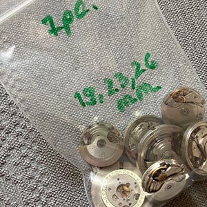 May include: A clear plastic bag containing several disassembled watch movements. The bag has green handwritten text that reads "7pc." and "19,23,26 mm." The watch parts are silver and have intricate internal mechanisms.