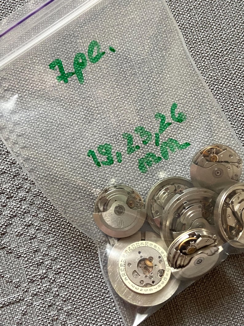 May include: A clear plastic bag containing several disassembled watch movements. The bag has green handwritten text that reads "7pc." and "19,23,26 mm." The watch parts are silver and have intricate internal mechanisms.