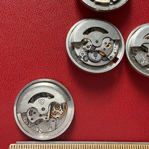 Puede incluir: Varios movimientos de reloj desmontados con engranajes y mecanismos intrincados visibles. Las piezas del reloj, de color plateado, est&aacute;n dispuestas sobre una superficie roja. Una regla de madera con marcas de mil&iacute;metros se encuentra en la parte inferior de la imagen.