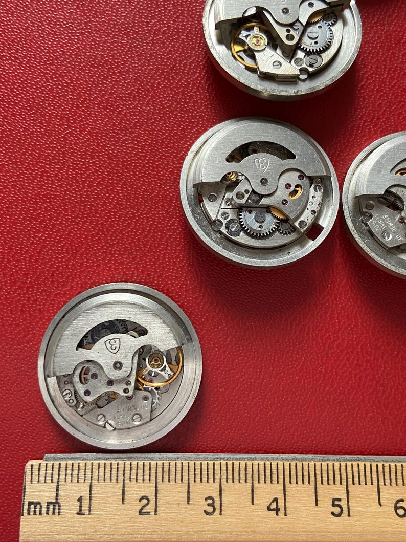 Puede incluir: Varios movimientos de reloj desmontados con engranajes y mecanismos intrincados visibles. Las piezas del reloj, de color plateado, est&aacute;n dispuestas sobre una superficie roja. Una regla de madera con marcas de mil&iacute;metros se encuentra en la parte inferior de la imagen.