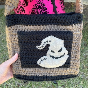 May include: A handmade crochet crossbody bag featuring a tan and black color scheme. The front panel displays a black square patch with a white, stitched ghost-like character appliqué. The bag includes a thick, textured crochet strap and trim.
