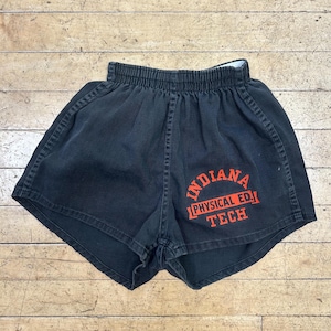 May include: Black athletic shorts with an elastic waistband and curved hem. The front features orange screen-printed text that reads INDIANA PHYSICAL ED. TECH in a stacked, arched layout with decorative brackets surrounding the middle line.