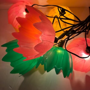May include: A string of lights with pink, green, and yellow plastic flower-shaped bulbs. The lights are connected by black wires.
