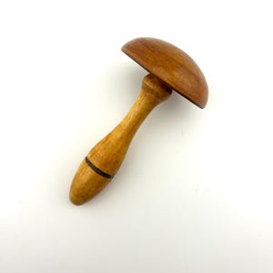 May include: A wooden darning mushroom with a smooth, light brown finish on the handle and a darker, rounded cap. The handle features a thin, dark horizontal ring detail near the base, showcasing a natural wood grain texture throughout the object.