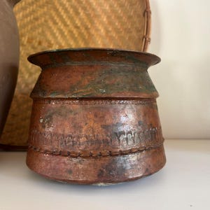 May include: A copper pot with a wide, flared rim and a tapered body. The surface features a weathered patina with green oxidation and horizontal bands of decorative etched patterns, including a row of small arches and a textured geometric border.