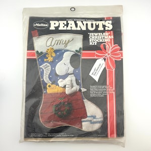 May include: A Malina Peanuts Jeweled Christmas Stocking Kit in original plastic packaging. The kit features a felt stocking design with Snoopy, Woodstock, and a Dear Santa letter. The packaging includes text, a red bow graphic, and a tag.