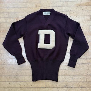 May include: A dark purple long-sleeve V-neck sweater featuring a large, cream-colored felt letter D on the chest. The garment has a wide ribbed hem and cuffs, and a white interior neck label that reads TOOL SHOP DETROIT.
