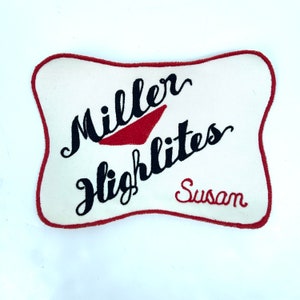 May include: White fabric patch with red stitching around the edges. The patch has black embroidered text that reads "Miller Highlife" and "Susan".