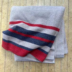 Peut inclure: Une couverture grise pli&eacute;e avec un motif &agrave; rayures audacieux en rouge, bleu marine et gris clair. La couverture est faite d'un mat&eacute;riau doux et est pli&eacute;e avec soin, mettant en valeur les rayures color&eacute;es.