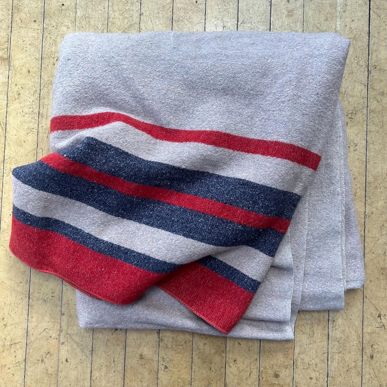 Peut inclure: Une couverture grise pli&eacute;e avec un motif &agrave; rayures audacieux en rouge, bleu marine et gris clair. La couverture est faite d'un mat&eacute;riau doux et est pli&eacute;e avec soin, mettant en valeur les rayures color&eacute;es.