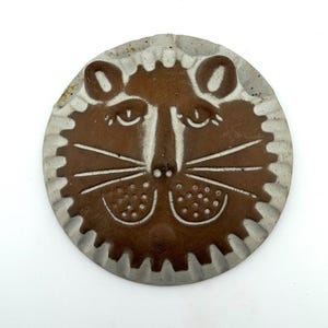 May include: A circular ceramic wall plaque featuring a stylized brown lion face with a cream-colored border. The lion has incised details including eyes, whiskers, and dotted muzzle areas, set against a smooth, flat, round clay surface.