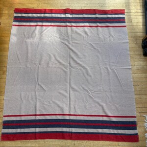 Peut inclure: Une grande couverture rectangulaire de couleur grise neutre. La couverture pr&eacute;sente des rayures rouges, bleu marine et blanches sur les bords sup&eacute;rieur et inf&eacute;rieur. La couverture est &eacute;tal&eacute;e sur un sol en bois.
