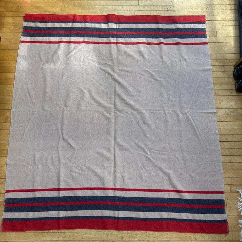 Peut inclure: Une grande couverture rectangulaire de couleur grise neutre. La couverture pr&eacute;sente des rayures rouges, bleu marine et blanches sur les bords sup&eacute;rieur et inf&eacute;rieur. La couverture est &eacute;tal&eacute;e sur un sol en bois.