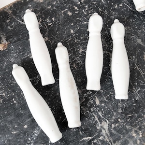 May include: A collection of white ceramic drawer pulls with a rounded top and a slender, elongated body. The pulls are arranged on a dark, textured surface, with a sprig of dried berries in the upper left corner.