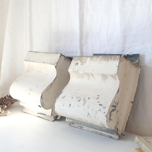 May include: Two vintage, white-painted wooden corbels with distressed paint, displayed on a white surface. The corbels feature a curved design and are likely architectural salvage. A small cluster of dried seed pods and a book are also visible.