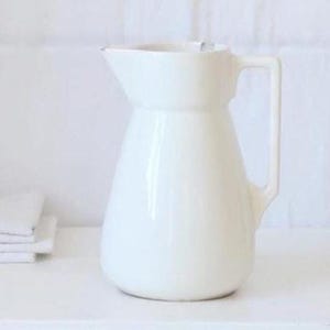 以下が含まれることがあります： 白い取っ手付きのセラミックピッチャー、折りたたまれた白いリネンの山、そして小さな丸い白い容器。これらのアイテムは白い背景に対して白い表面に配置されており、ミニマリストな美学を作り出しています。