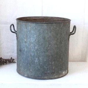 May include: A weathered, cylindrical metal bucket with two curved handles. The bucket has a distressed, gray finish and a slightly rusted rim. A small bundle of dried flowers rests nearby, suggesting a rustic, farmhouse aesthetic.