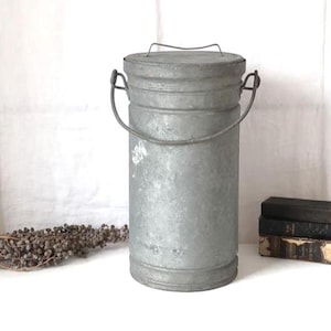 May include: A vintage galvanised metal milk churn with a lid and a handle. The churn is cylindrical and has a distressed, aged appearance. A small bundle of dried flowers and a stack of books are nearby.