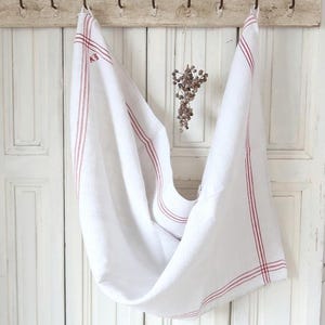 以下が含まれることがあります： 赤色のストライプが入った白いリネンのタオルが、古びた白いドアの素朴な木製ラックに掛けられています。タオルの間には、小さなドライフラワーの束が吊り下げられています。下には白く古びたテーブルがあります。