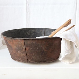 Op de afbeelding: Een ronde, donkere, roestige metalen kom met een verweerd oppervlak. Een witte doek hangt over de rand en een houten gereedschap ligt in de kom. Een stapel van vier gevouwen witte doeken ligt links van de kom op een vlakke ondergrond.