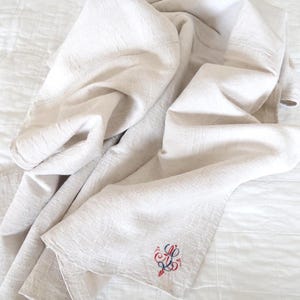 Può includere: Una coperta di lino color crema con una trama strutturata è drappeggiata su una superficie trapuntata bianca. La coperta ha un disegno ricamato in rosso, blu e bianco nell'angolo. La coperta è piegata e drappeggiata, mostrando la sua consistenza e il suo design.