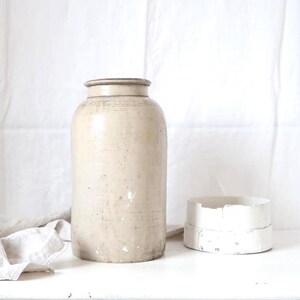 Puede incluir: Una vasija de gres alta y cilíndrica en color beige claro con un borde liso y una textura de superficie desgastada y moteada. A su lado se encuentra un pequeño recipiente circular de madera blanca con un borde ligeramente astillado.