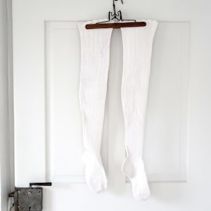 Op de afbeelding: Een paar witte kniehoge sokken hangen aan een donkerbruine hanger aan een witte deur. De sokken zijn gemaakt van een dik, getextureerd gebreid materiaal. De deur heeft een vintage metalen slot.