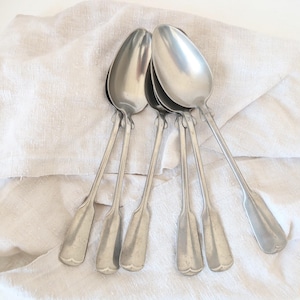 May include: Six vintage silver spoons are displayed on a cream-coloured linen cloth. The spoons have a classic design with elongated bowls and decorative handles. These spoons are likely for serving or eating.