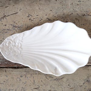 Op de afbeelding: Een witte keramische serveerschaal in de vorm van een schelp met een reliëf bloemmotief.