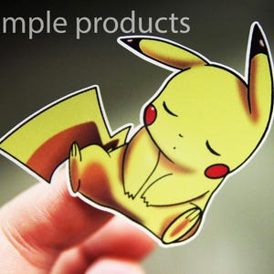May include: A yellow cartoon character with closed eyes and red cheeks. The character is a Pikachu from the Pokemon franchise. The character is printed on a sticker.