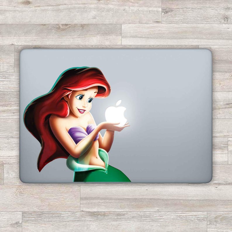May include: A laptop sticker featuring a cartoon image of Ariel, the Little Mermaid, holding a glowing white apple. Ariel is wearing a green tail and a purple seashell bra. Her hair is red and flowing.