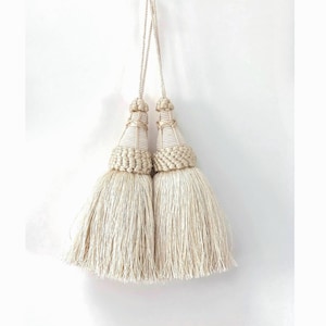 May include: A pair of off-white decorative tassels hanging from a single cord. Each tassel features a structured, wrapped conical top with a braided collar and a long, dense fringe skirt made of fine, silky threads that hang straight down.