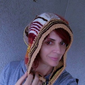 Può includere: Una donna indossa un cappello di lana a righe marroni, bianche e rosse con un lungo cordoncino all'uncinetto marrone, bianco e giallo.