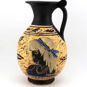 May include: A ceramic pitcher featuring a black neck and handle with a cream-colored body. The body displays a black and blue profile illustration of a person with long hair, accented by brown mottled textures and black leaf-like patterns.