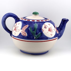 May include: A ceramic teapot featuring a hand-painted design of white fish with red fins and green seaweed on a deep blue background. The lower portion of the teapot is cream-colored with a thin red horizontal stripe, and it includes a matching lid.