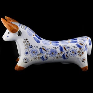 May include: A ceramic bull figurine featuring a white glazed surface decorated with hand-painted blue floral patterns and brown accents on the horns and hooves. The bull is depicted in a standing position with detailed blue petals and leaves.