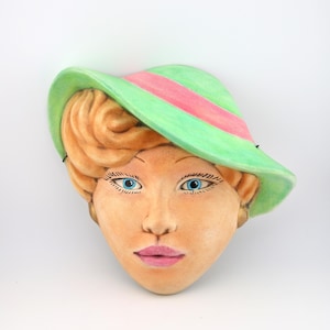 May include: A ceramic wall mask depicting a face with blonde hair, bright blue eyes, and pink lips. The figure wears a wide-brimmed light green hat featuring a prominent pink band. The skin tone is a warm tan, and the facial features are painted.