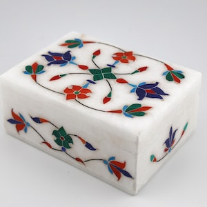 May include: A rectangular white marble box featuring an inlaid floral pattern on the lid and front side. The design consists of stylized flowers and leaves in vibrant shades of red, blue, and green, creating a symmetrical decorative motif.