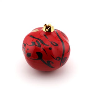 May include: A glossy red ceramic pomegranate sculpture featuring black calligraphic script wrapping around its rounded surface. The top of the fruit is accented with a metallic gold crown-like stem, providing a contrast to the vibrant red finish.